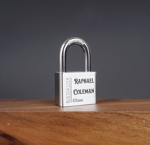 A custom stainless steel padlock with the name 'Raphael Coleman' engraved on the shackle, displayed on a wooden surface.